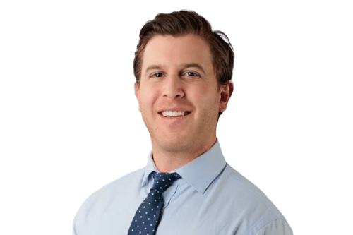 Portrait of a smiling man in a light blue dress shirt and navy polka-dot tie against a solid background, looking at the camera.