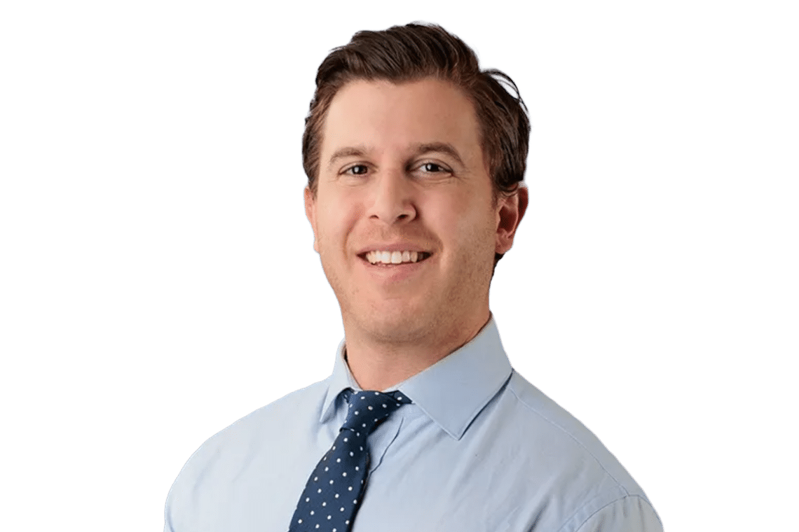 Portrait of a smiling man in a light blue dress shirt and navy polka-dot tie against a solid background, looking at the camera.
